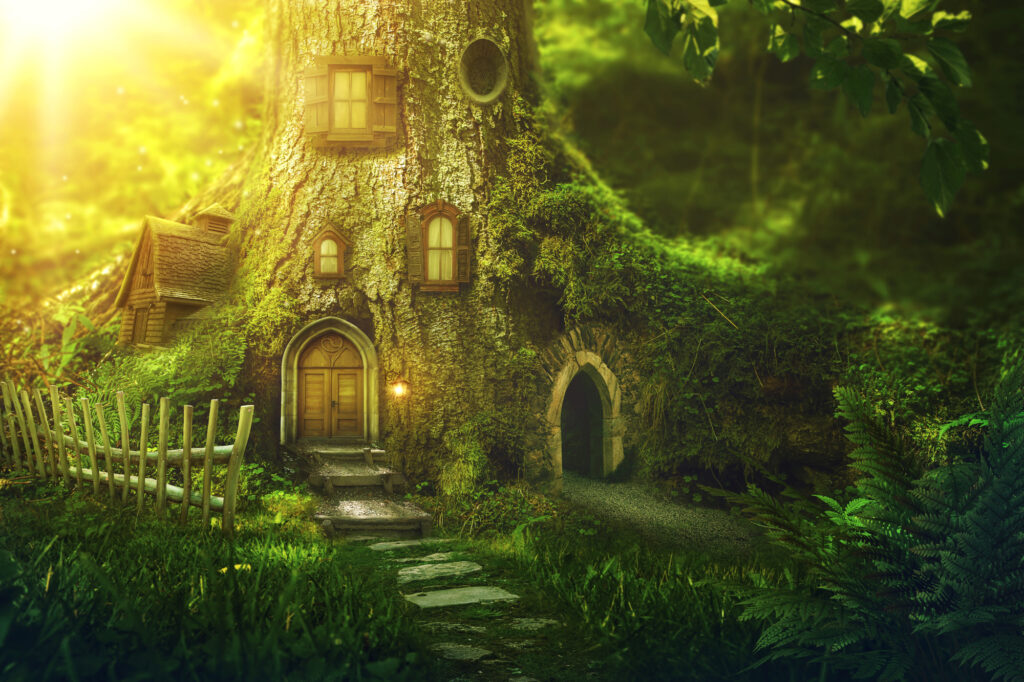 Ein feen- oder wichtelhaftes Baumhaus in einer grünen Waldbodenlandschaft mit Moos. Das Haus befindet sich im Stamm eines alten Baums, er hat deshalb Fensterchen und kleine Türen. Eine Treppe führt zum Eingang hinauf. Märchenhafter Eindruck.
